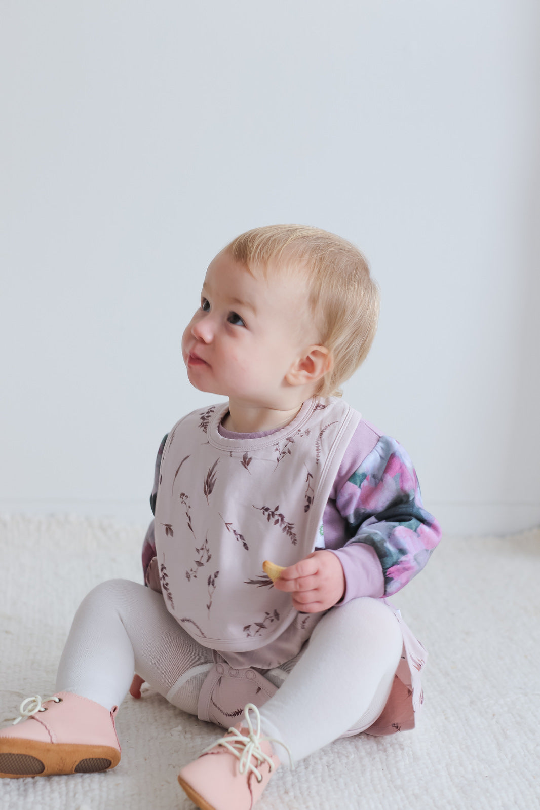 Girl in Violet Long Sleeve Sweater, Snuggle Bib, Wanders Shoe and Knitted Footed Tights in White, all designed by Burrow and Be