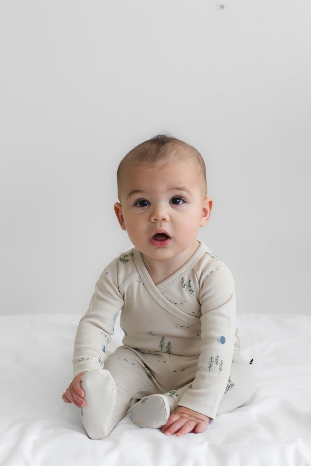 Baby sitting on white sheets in front of a white wall, wearing Midnight Forest Footed Pants, designed by Burrow and Be