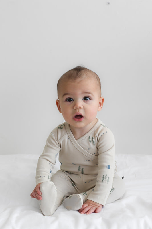 Baby sitting on white sheets in front of a white wall, wearing Midnight Forest Footed Pants, designed by Burrow and Be