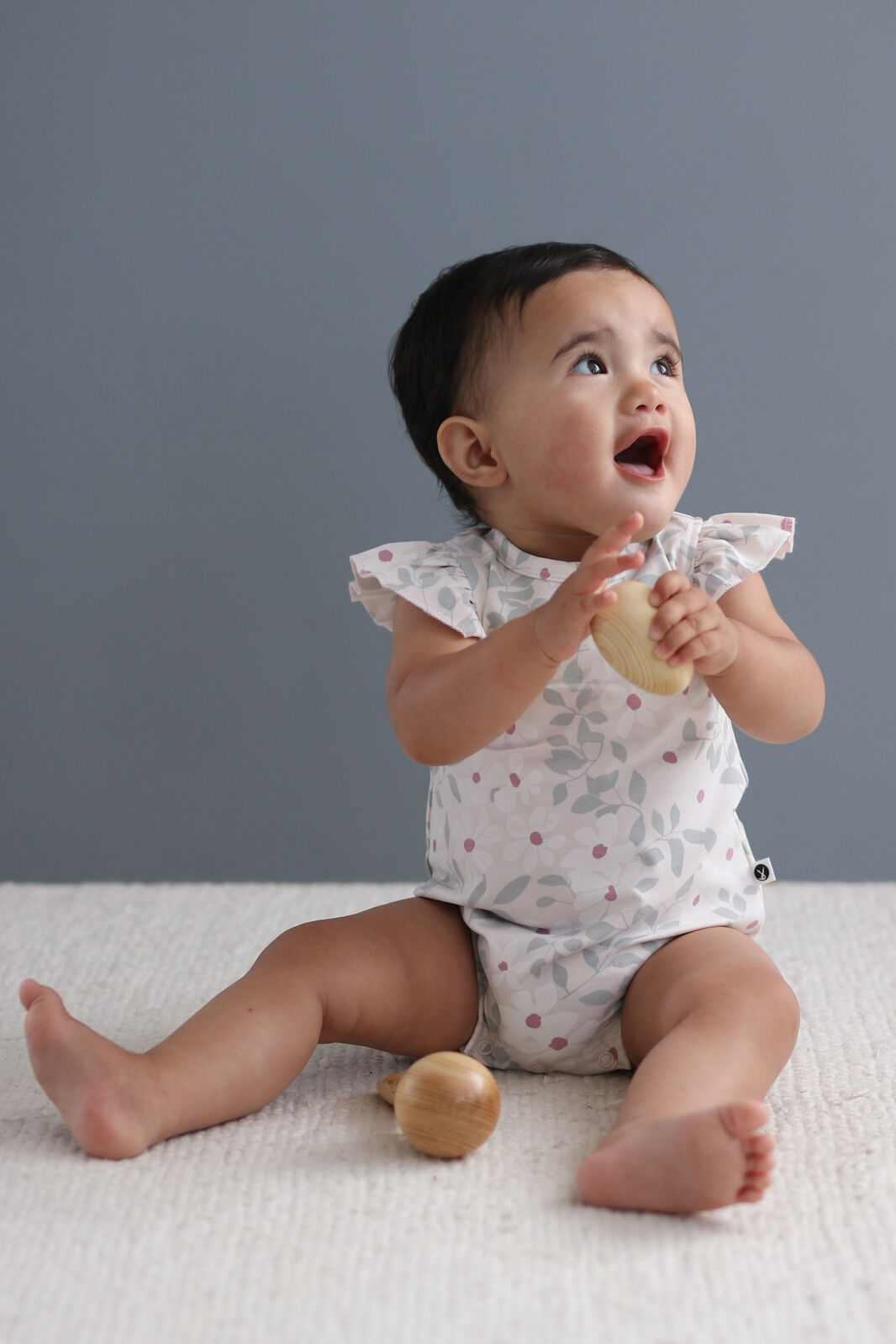 Baby sitting on a white surface with wooden toys, wearing a patterned onesie against a gray background.