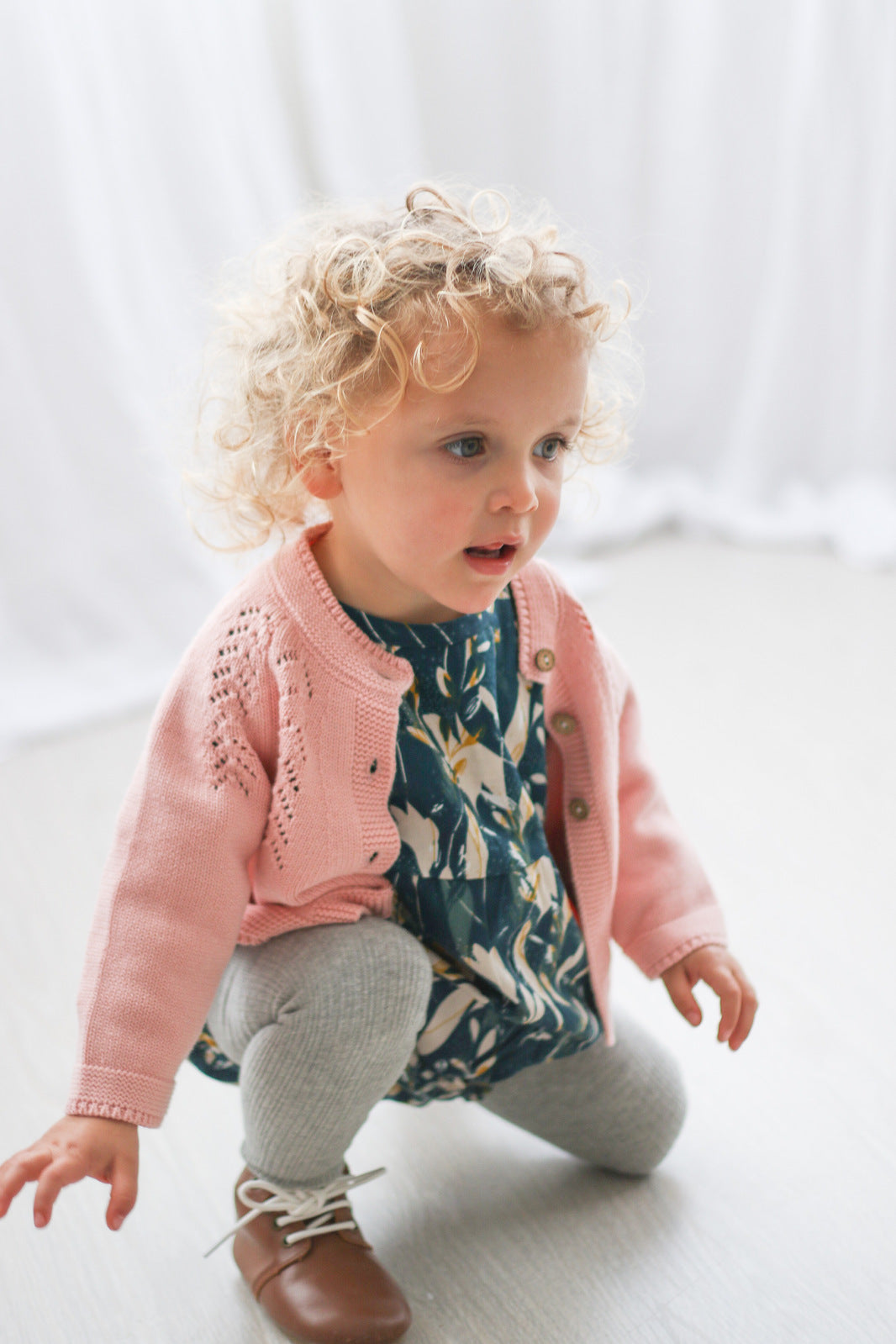 Girl kneeling on wooden floor in front of a white curtain, wearing Green Leavings Top with Grey Knitted Footed Tights, Shoes by Wanders and Lily Knit Cardigan, all designed by Burrow and Be