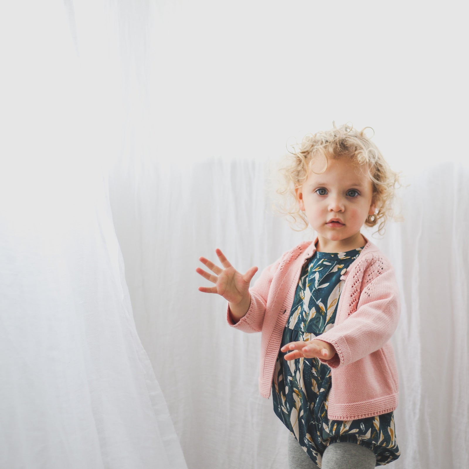 Girl standing in front of a white curtain, wearing Green Leavings Bubble Romper with Grey Knitted Footed Tights and Lily Knit Cardigan, all designed by Burrow and Be