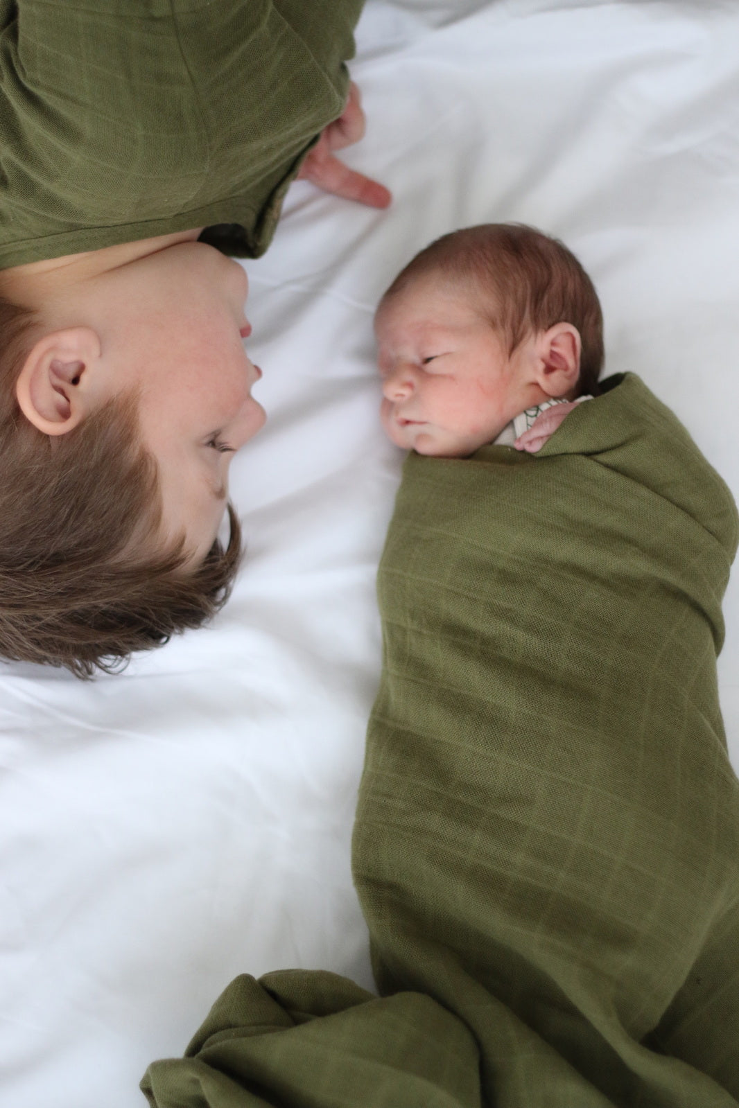 Boy wearing a a Pine Jasper woven top lying on the bed with Baby wrapped in Essential Muslin Wrap in Pine, both designed by Burrow and Be