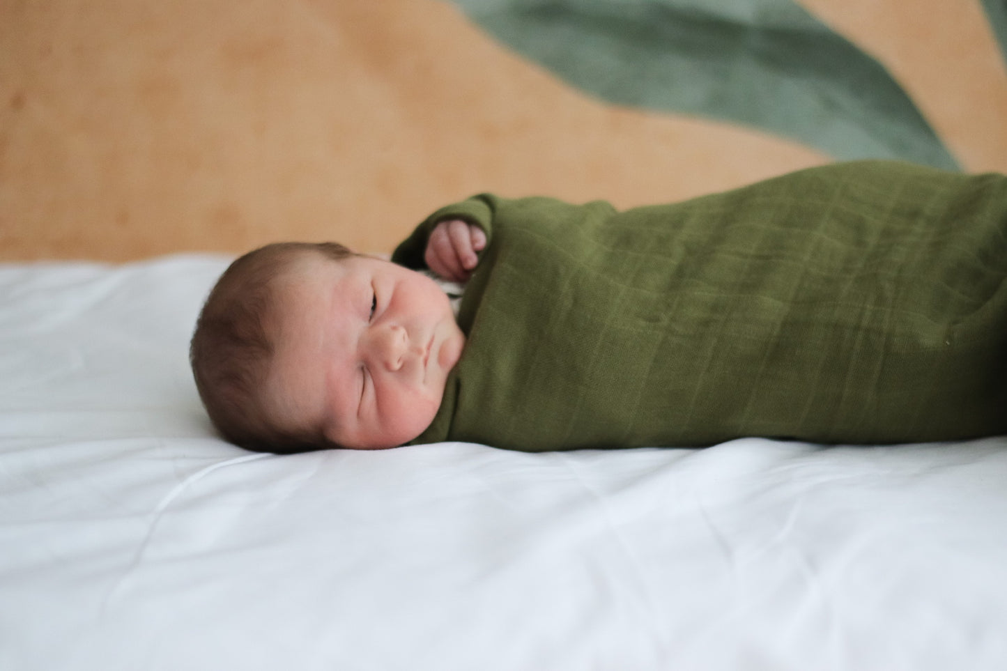 Baby lying on white sheets, wrapped in Essential Muslin Wrap in Pine, designed by Burrow and Be