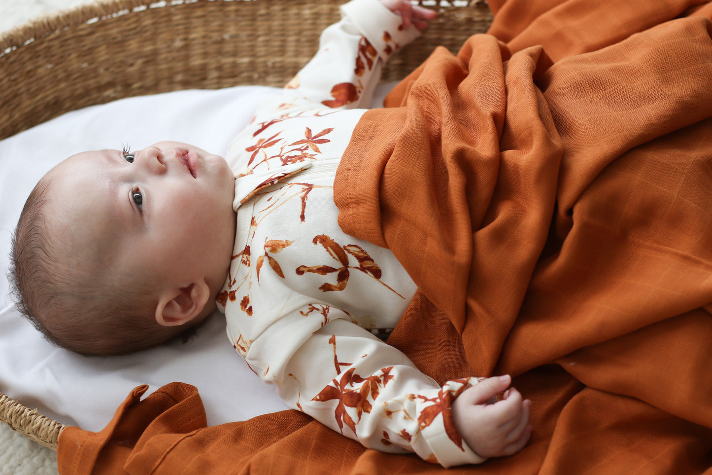 Baby in a Moses Basket wearing Autumn Leaves Zip Suit and covered with Essential Muslin Wrap in Clay, designed by Burrow and Be