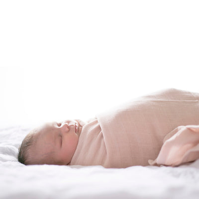 Baby lying on white sheets wrapped in Essential Muslin Wrap in Blush, designed by Burrow and Be