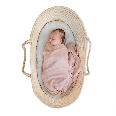 Baby in a Moses basket, wrapped in Essential Muslin Wrap in Dusty Rose, designed by Burrow and Be