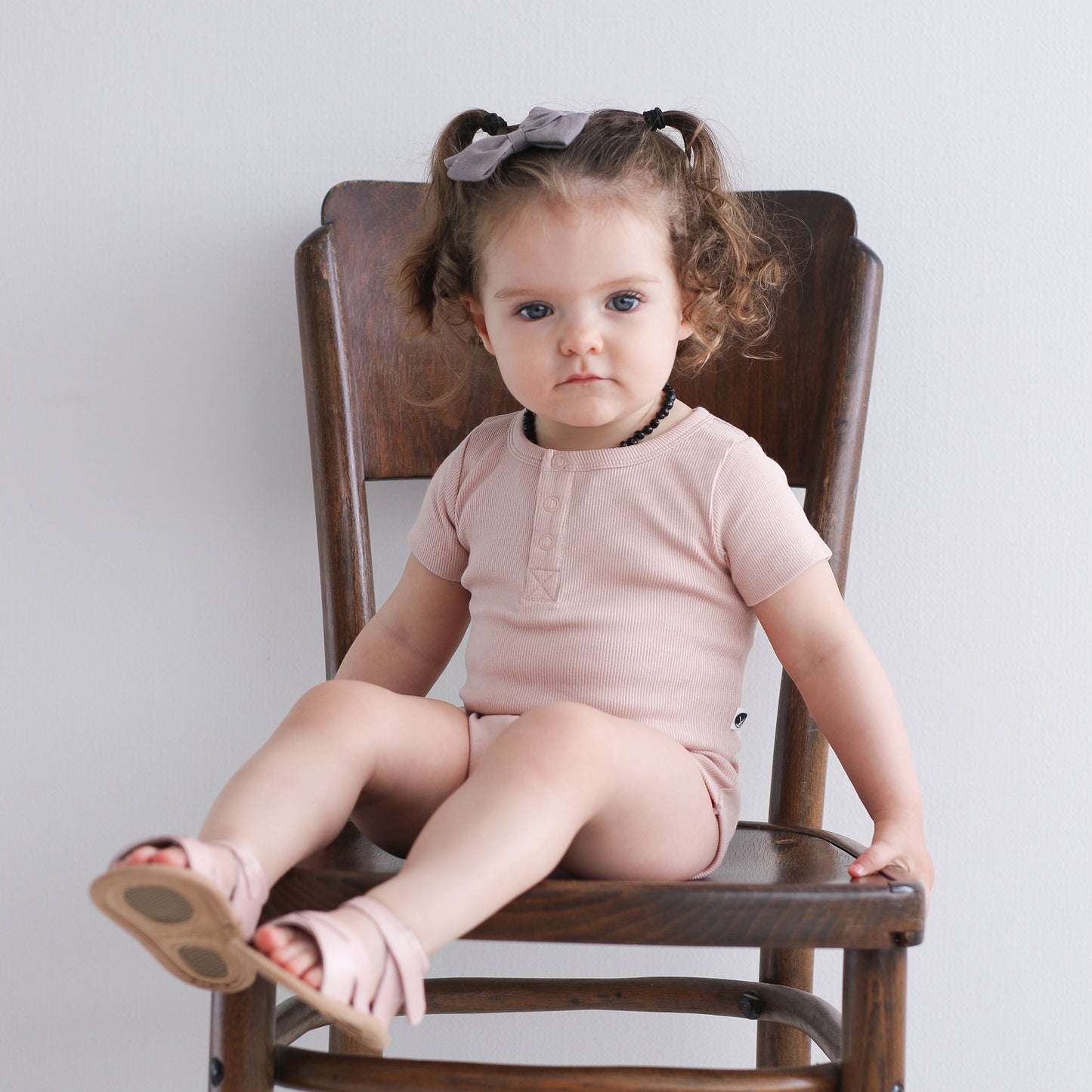 Baby sitting on wooden chair in front of a white wall, wearing Sandals by Wander and Short Sleeve Henley Rib Bodysuit in Dusty Rose, all designed by Burrow and Be