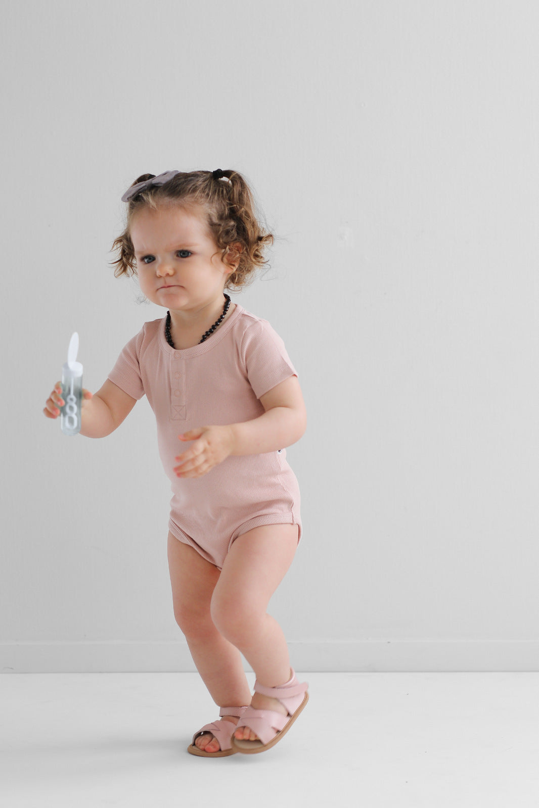 Baby playing with bubbles in front of a white wall, wearing Sandals by Wander and Short Sleeve Henley Rib Bodysuit in Dusty Rose, all designed by Burrow and Be