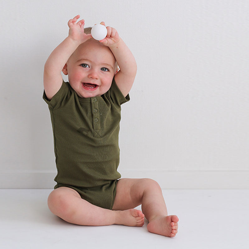 Baby sitting on floor in front of a white wall, holding a ball and wearing a Short Sleeve Henley Rib Bodysuit in Olive, designed by Burrow and Be