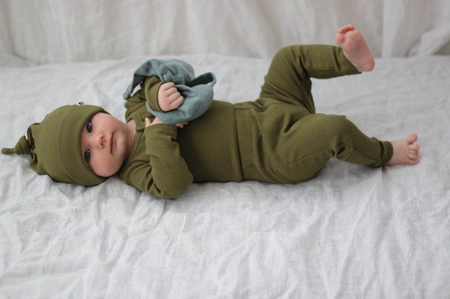 Baby lying on the bed wear Beanie, Bodysuit and Essential Derby Rib Leggings, all in Olive colour and designed by Burrow and Be