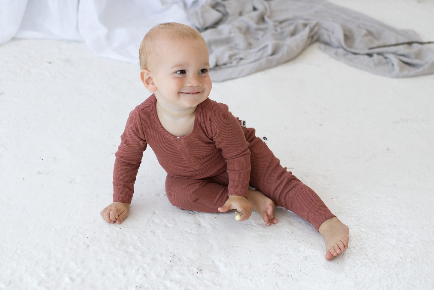 Baby posing and smiling at the camera while wearing Long Sleeve Bodysuit and Essential Derby Rib Leggings, all in Clay and designed by Burrow and Be