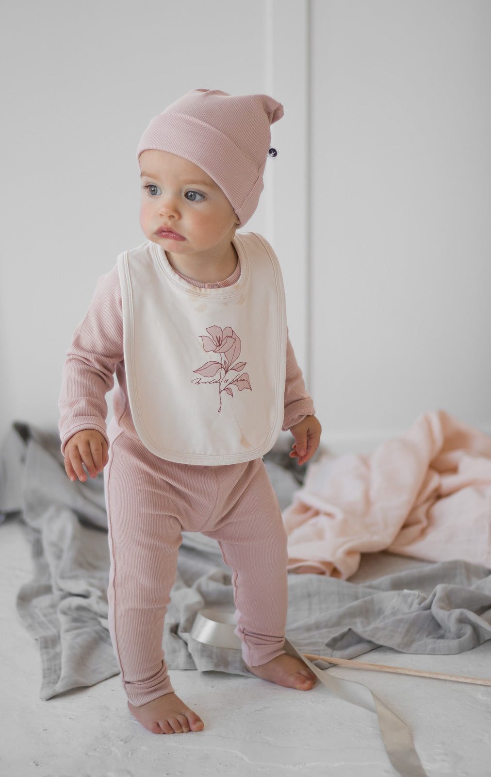 Baby wearing Wildflower Snuggle Bib, Beanie, Bodysuit and Essential Derby Rib Leggings, all in Dusty Rose and designed by Burrow and Be