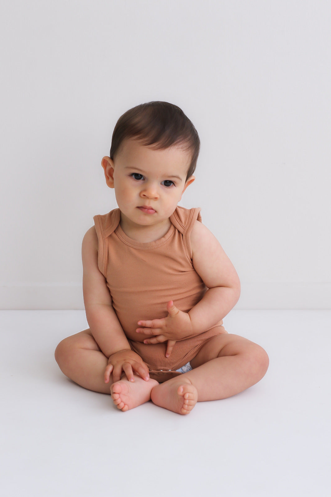 Baby boy sitting against a white background wearing Essential Singlet Bodysuit in Tawny Brown, designed by Burrow and Be