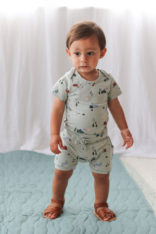 Baby boy wearing Shoes by Wanders, Garden Treasures Shorts and Garden Treasures Short Sleeve Bodysuit against white curtains and standing on Sage Quilted Mat, all designed by Burrow and Be