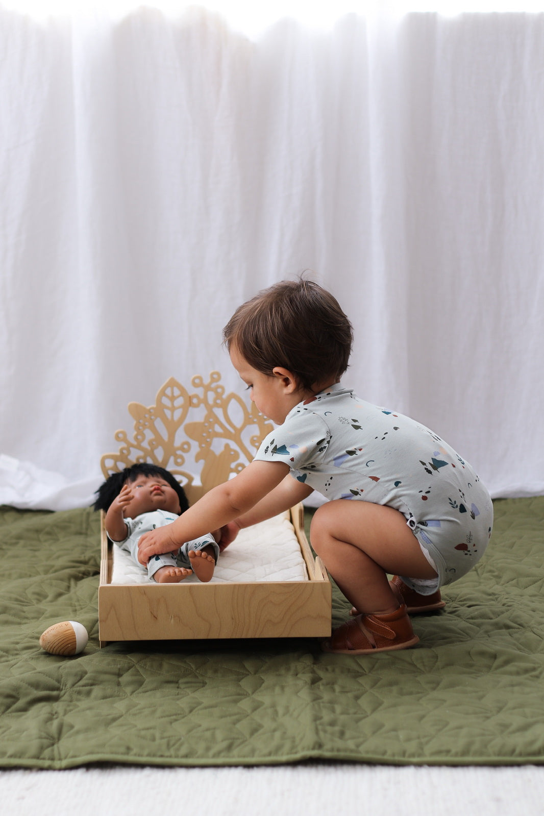 Baby boy playing with a doll dressed in Garden Treasure while wearing Shoes by Wanders, Garden Treasures Shorts and Garden Treasures Short Sleeve Bodysuit against white curtains and standing on Olive Quilted Mat, all designed by Burrow and Be