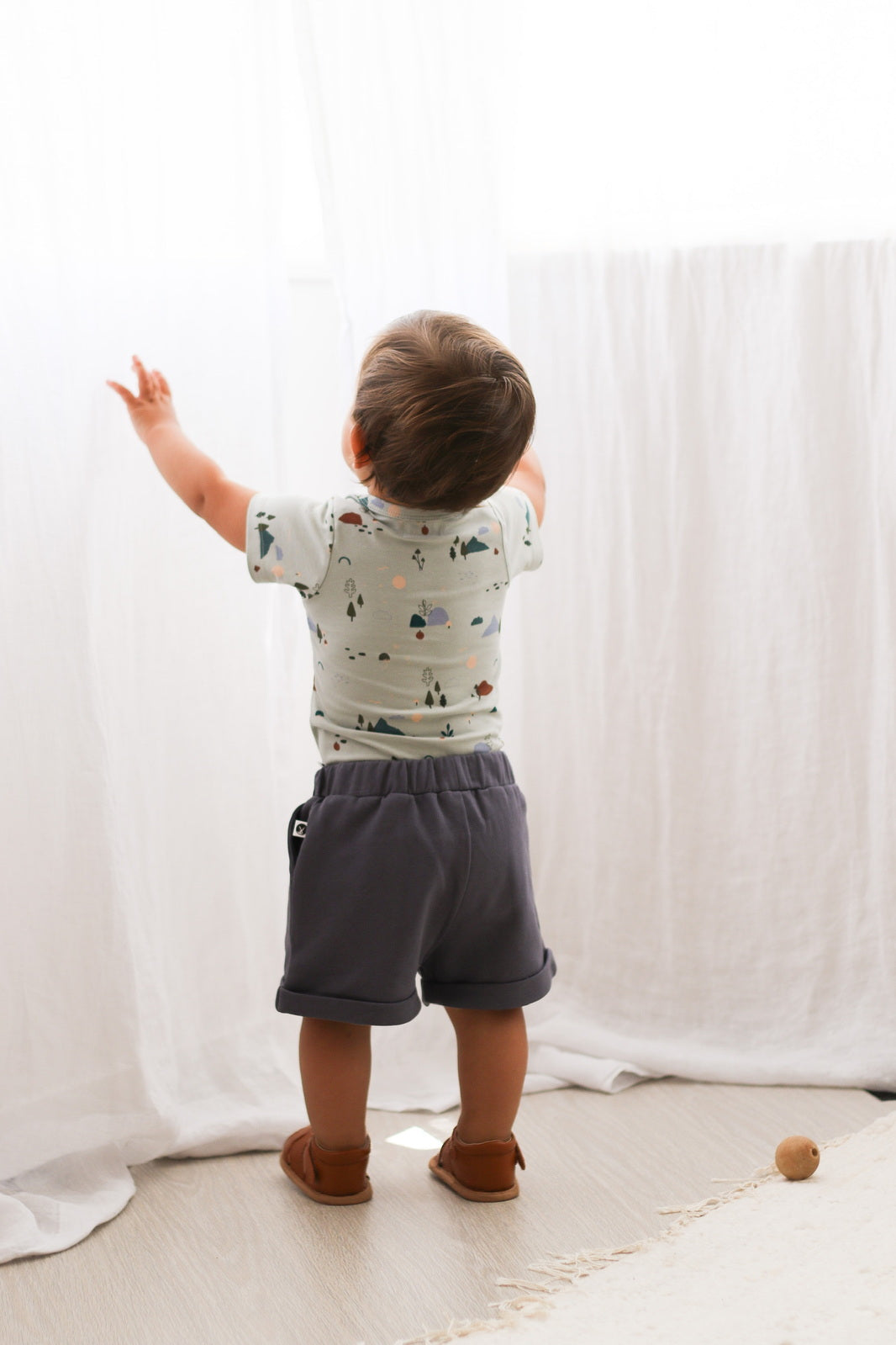 Baby boy wearing Shoes by Wanders, Ink Blue Terry Knit Baby Shorts and Garden Treasures Short Sleeve Bodysuit against white curtains, all designed by Burrow and Be