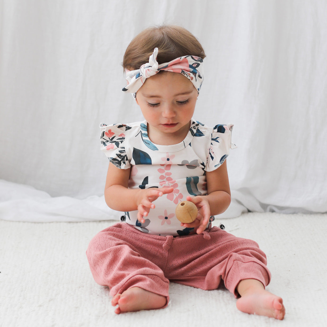Baby sitting on white carpet in front of a white curtain wearing Pointelle Pants in Dusk, Pink Clementine Headband and Flutter Bodysuit, all designed by Burrow and Be
