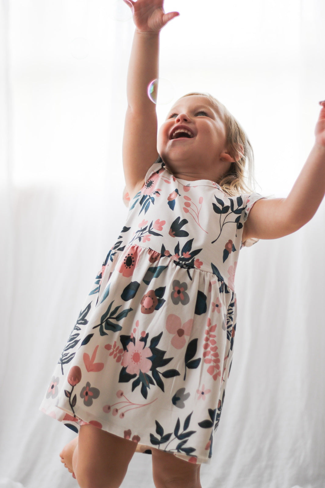 Girl playing with bubbles while standing in front of white curtains and wearing Pink Clementine Flutter Dress, designed by Burrow and Be