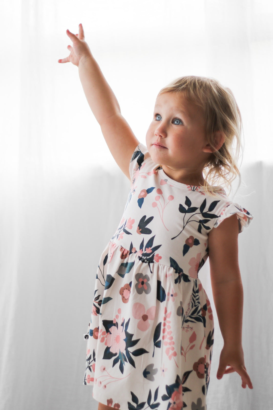 Girl standing in front of white curtains and wearing Pink Clementine Flutter Dress, designed by Burrow and Be