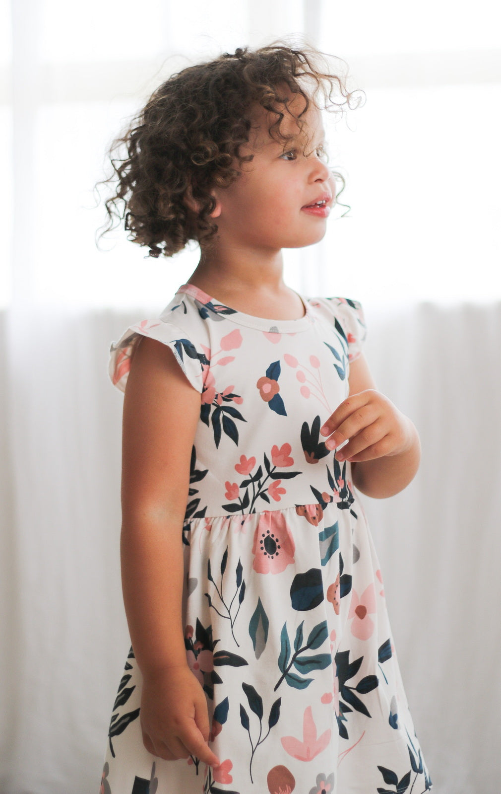 Girl standing in front of white curtains and wearing Pink Clementine Flutter Dress, designed by Burrow and Be