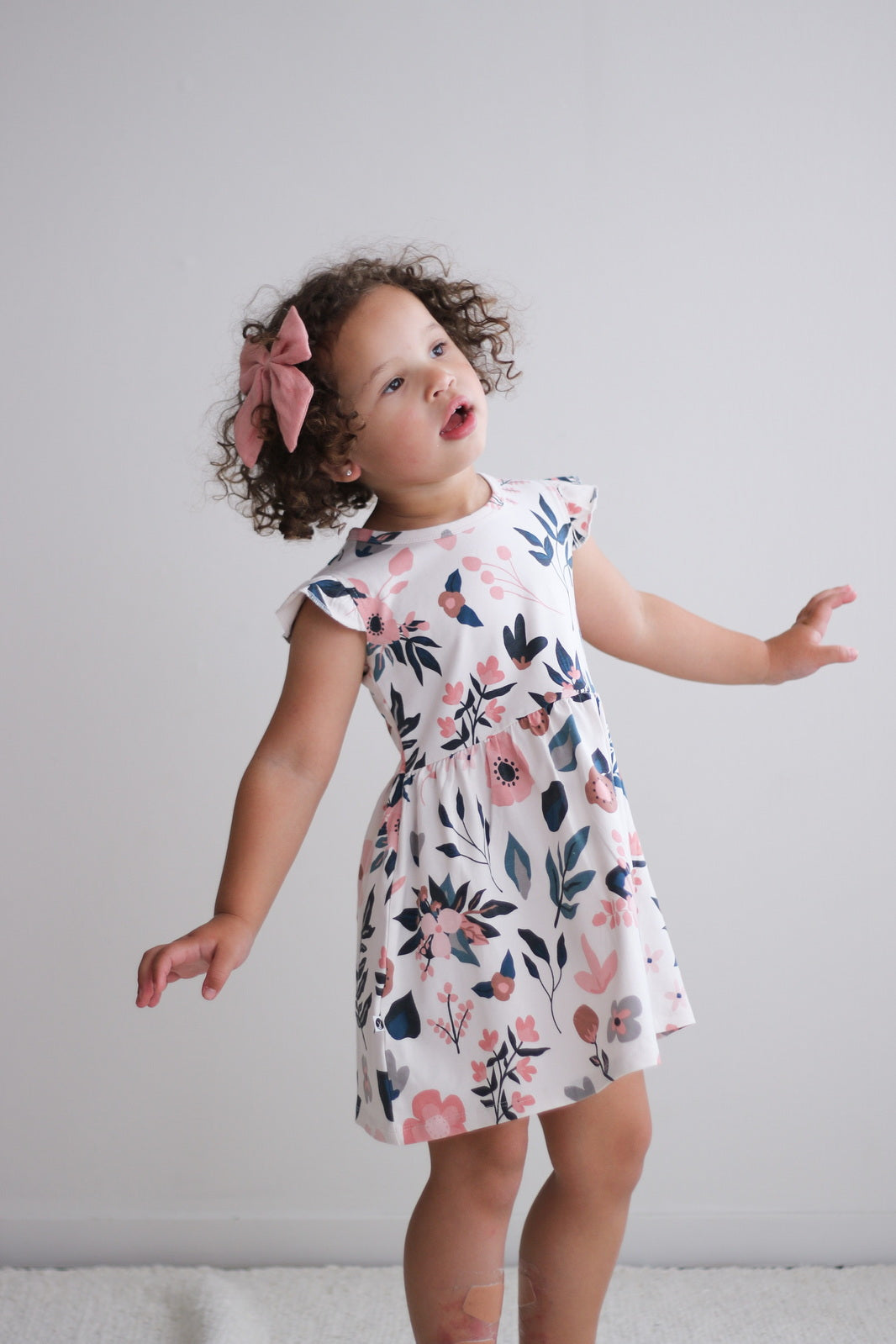 Girl standing in front of white wall and wearing Pink Clementine Flutter Dress, designed by Burrow and Be