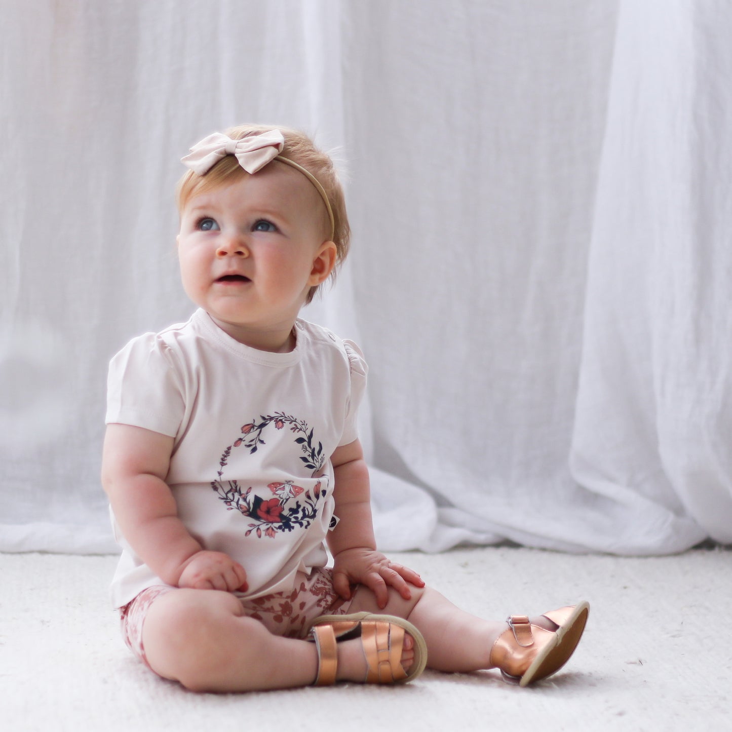 Baby girl sitting on carpet, wearing Fairy Wreath T-shirt, Flower Splash Bike shorts and Wanders Sandals, all designed by Burrow and be
