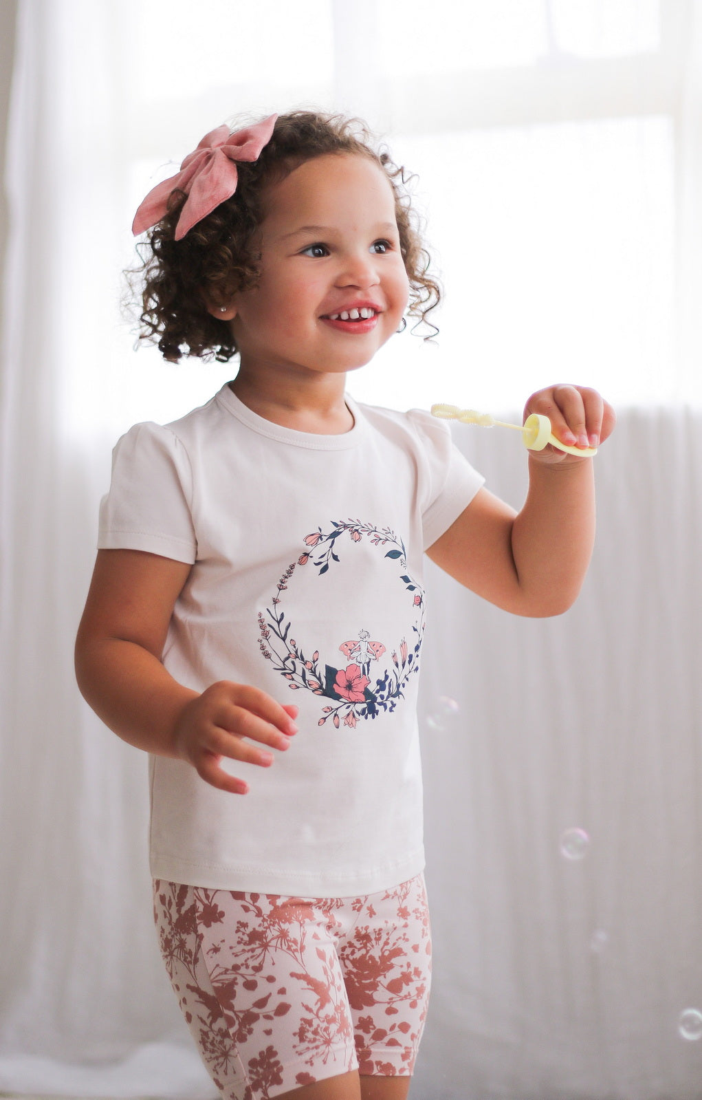Girl posing against white curtains while wearing Fairy Wreath T-shirt and Flower Splash Bike Shorts, both designed by Burrow and be