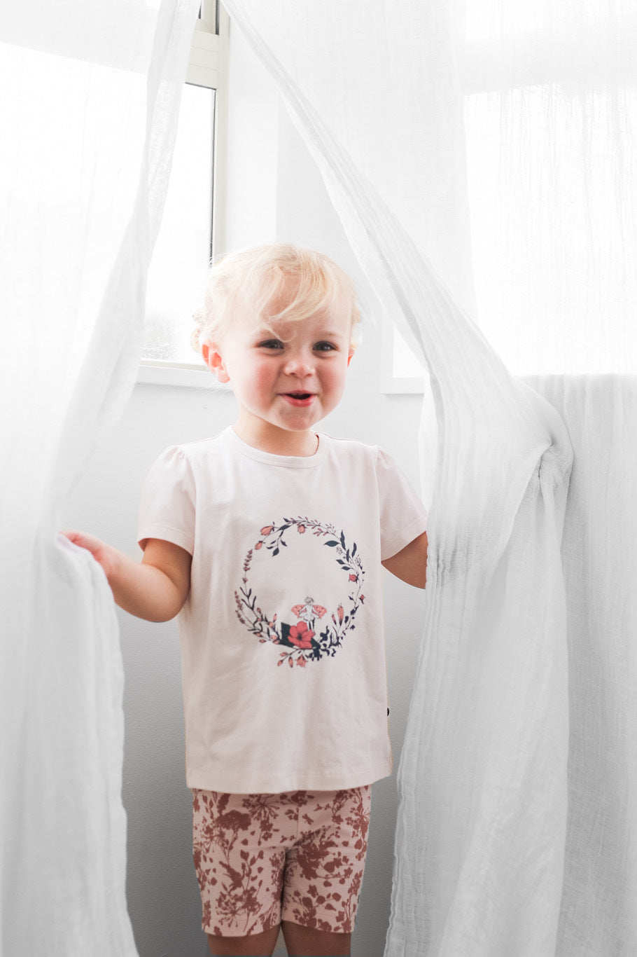 Girl standing in front of window while holding white curtains and wearing Fairy Wreath T-shirt and Flower Splash Bike Shorts, all designed by Burrow and be