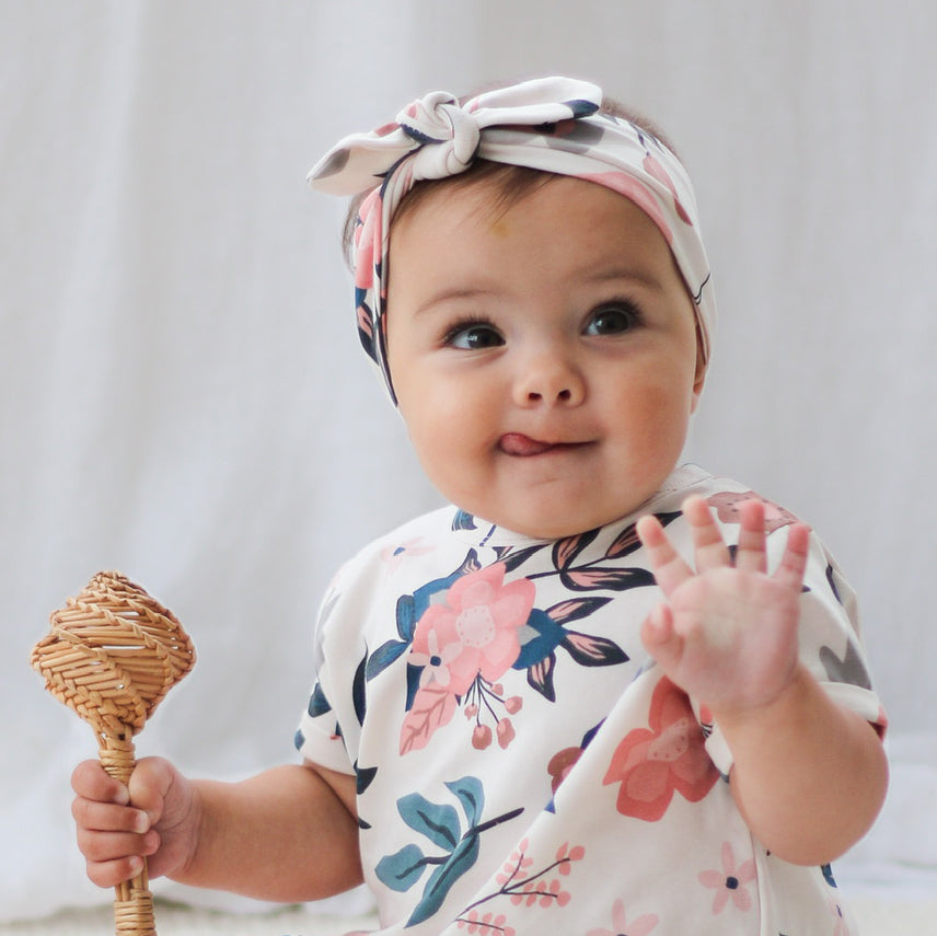 Baby girl holding wooden rattan toy while wearing T-shirt and Essential Baby Headband both in Pink Clementine print, designed by Burrow and Be