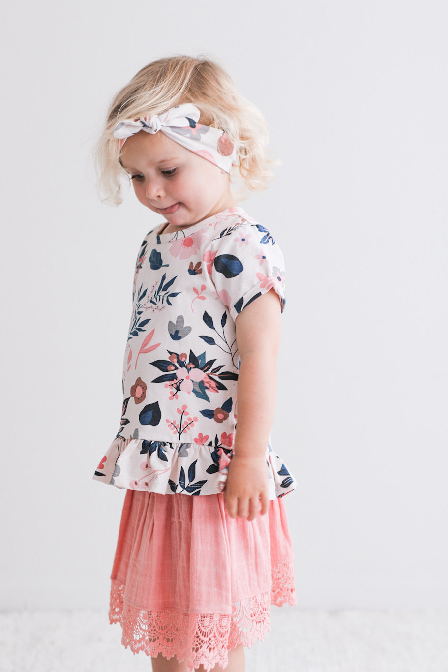 Baby girl looking down and posing while wearing Hana Top and Essential Baby Headband both in Pink Clementine print, designed by Burrow and Be