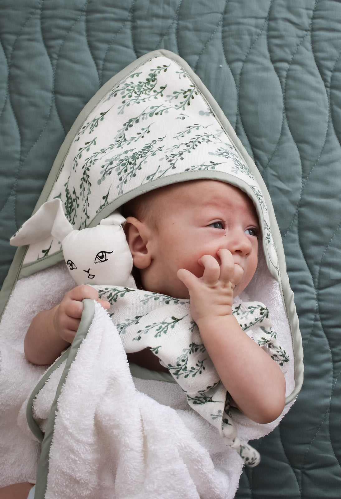 Baby laying on Essential Quilted Mat in Sage and wrapped in String of Pearls Baby Hooded Towel, both designed by Burrow and Be