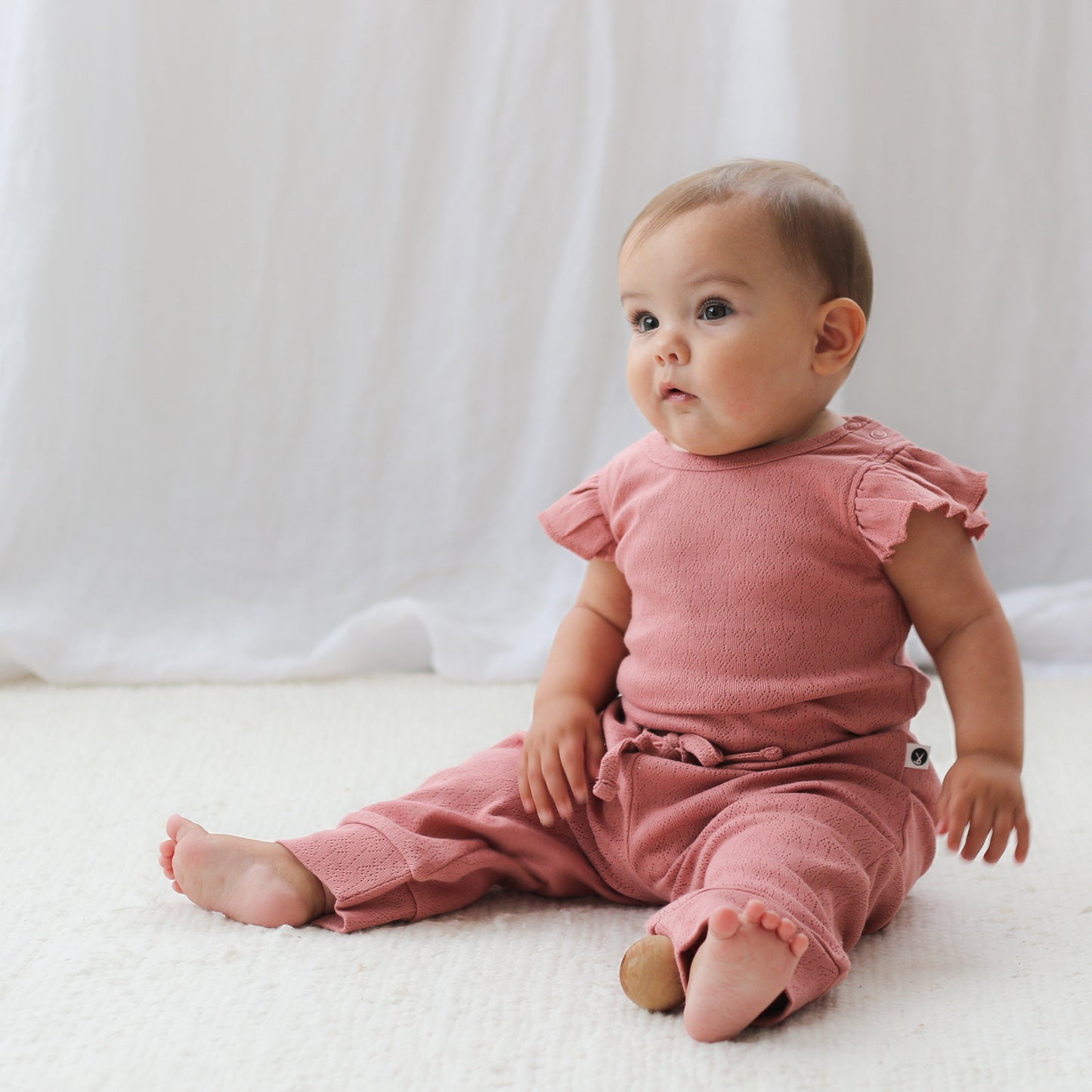 Baby girl sitting on carpet, wearing Pointelle Flutter Sleeve Bodysuit and Baby Pants in Dusk, both designed by Burrow and Be