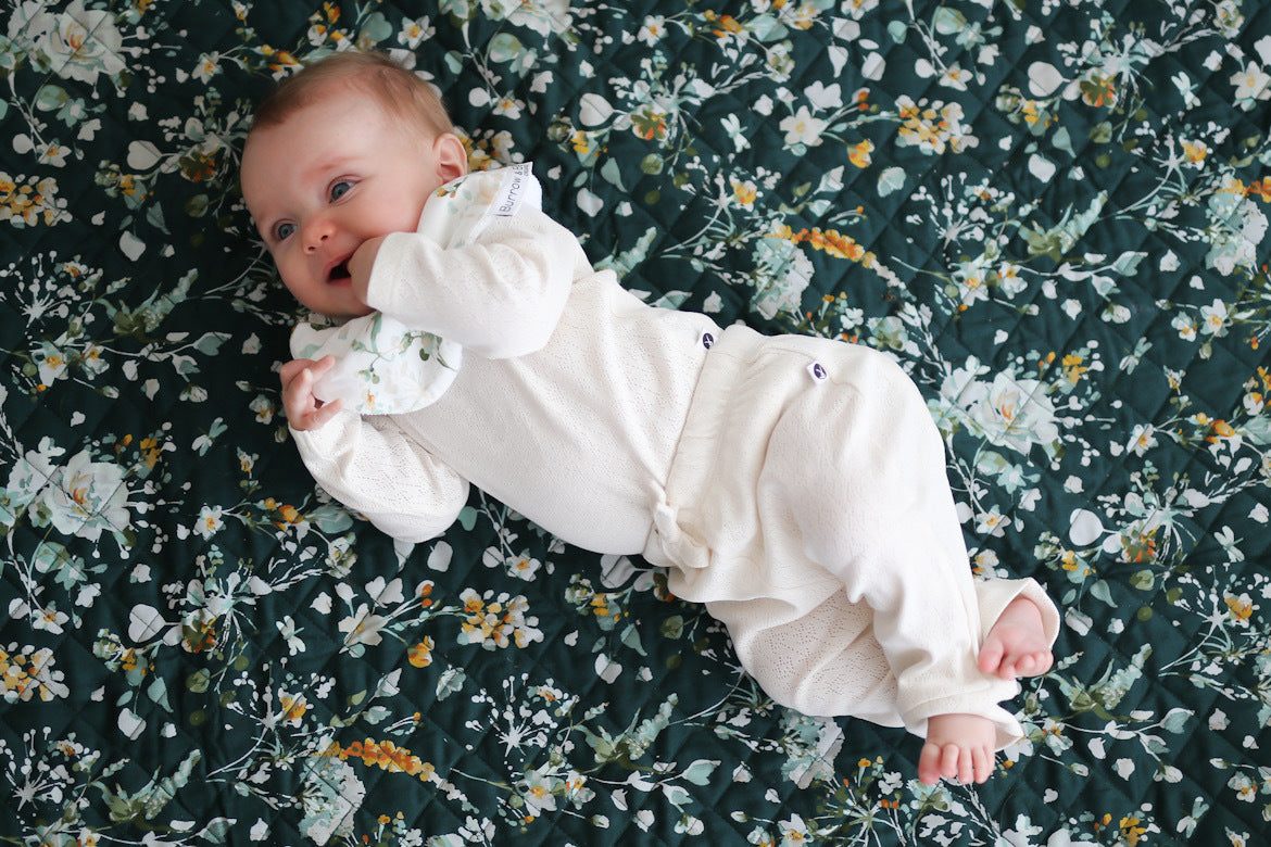 Baby laying on Green Leavings Quilted Mat while wearing Leavings Dribble Bib, Pointelle Long Sleeve Bodysuit and Baby Pants in Natural, all designed by Burrow and Be