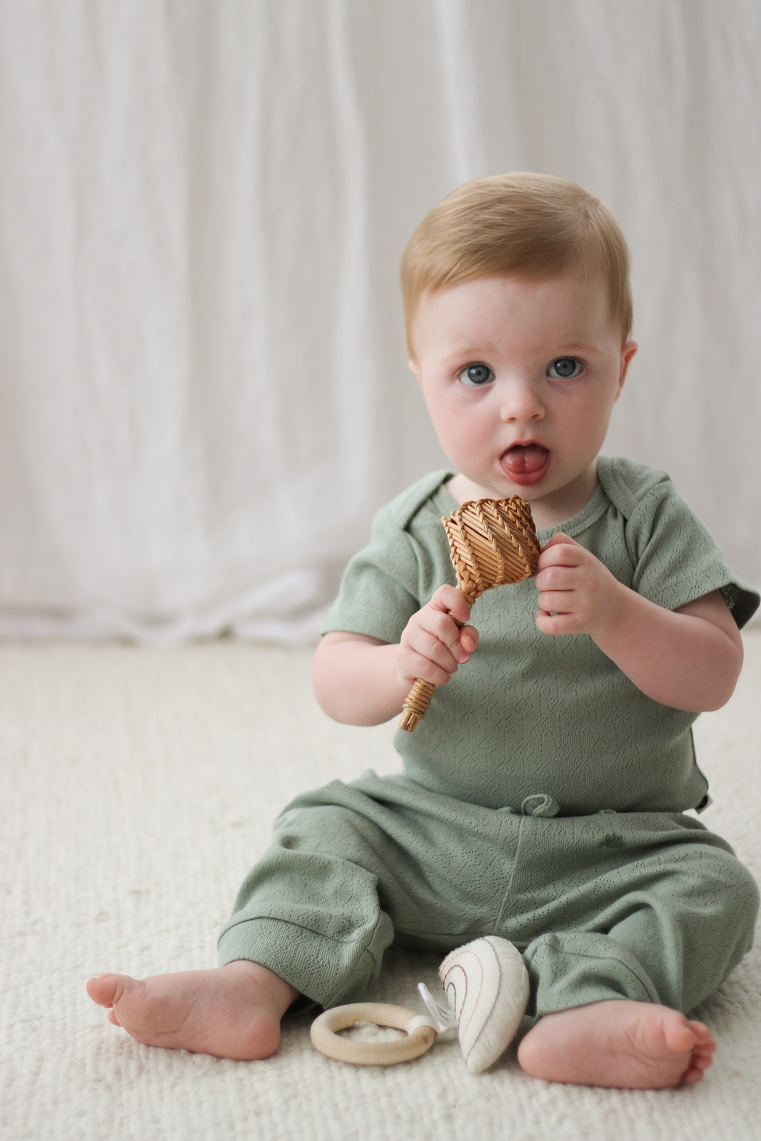 Baby sitting on carpet, in front on white curtains while wearing Pointelle Short Sleeve Bodysuit and Baby Pants in Riverstone, all designed by Burrow and Be