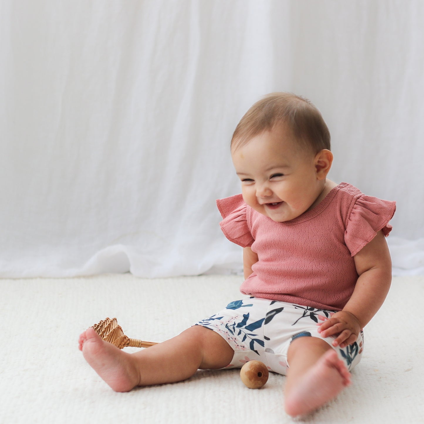 Baby sitting on wooden floor in front of white curtain, smiling and wearing Pink Clementine Bike Shorts and Pointelle Flutter Sleeve Bodysuit in Dusk, all designed by Burrow and Be