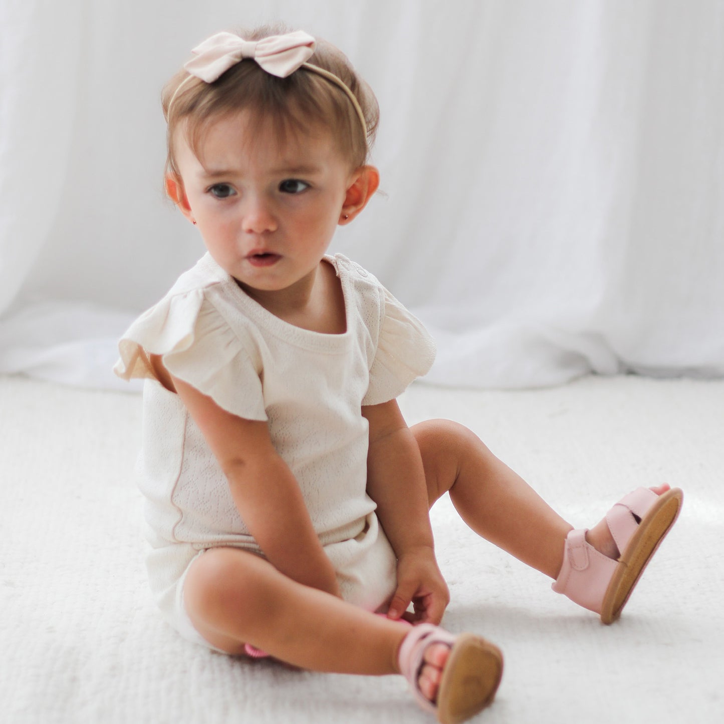 Baby girl sitting on white carpet and wearing Pointelle Flutter Sleeve Bodysuit and Shorts in Natural, all designed by Burrow and Be