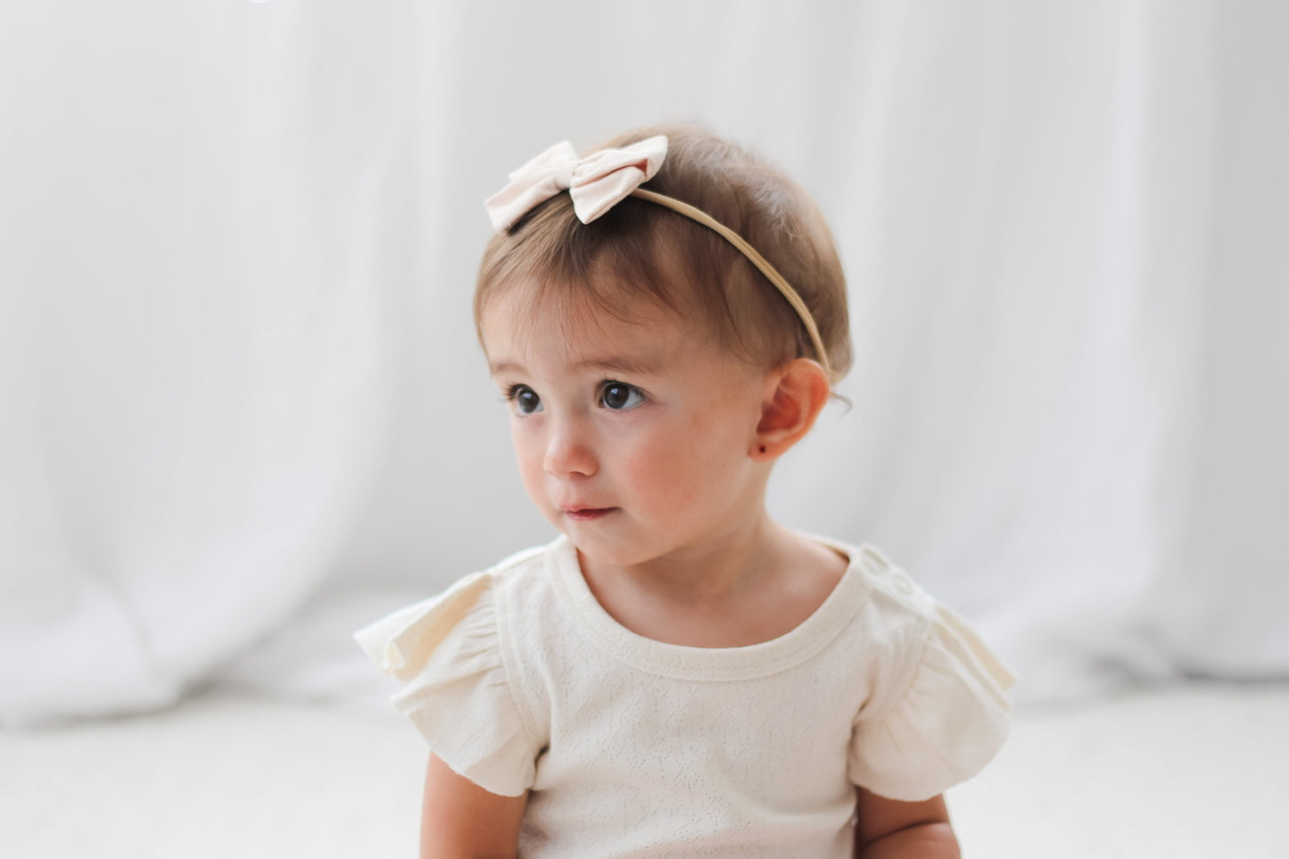 Baby girl sitting in front of white curtains, wearing Bow Tie Hair Tie and Pointelle Flutter Sleeve Bodysuit in Natural, all designed by Burrow and Be