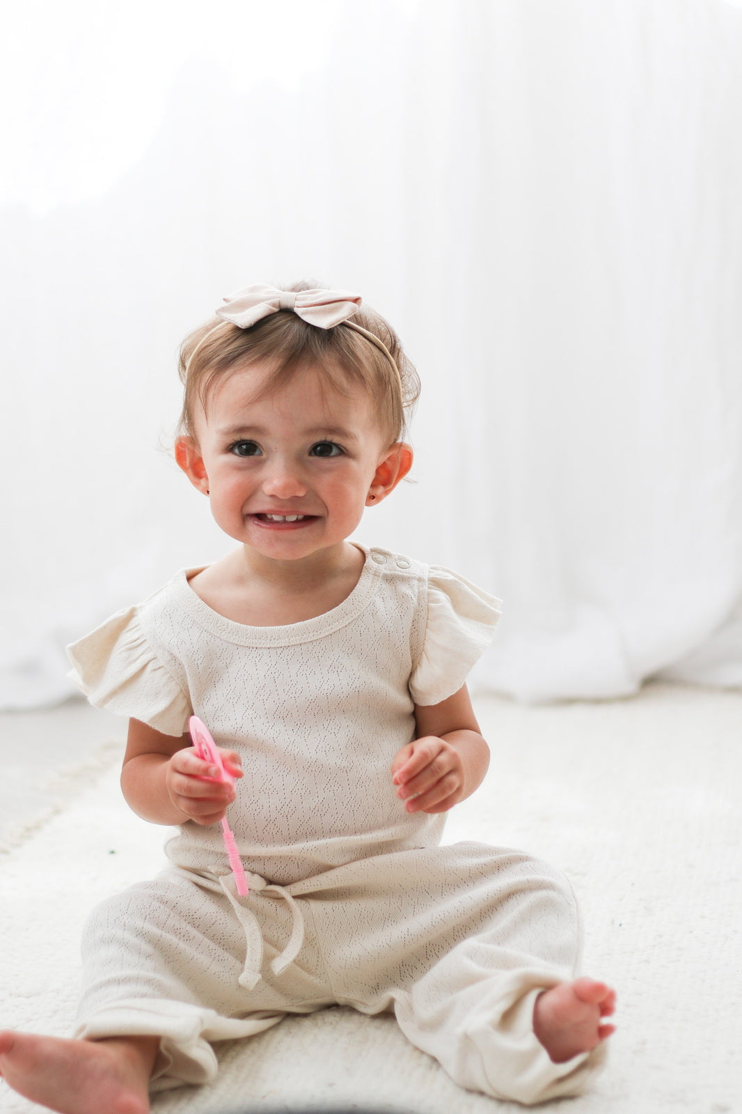 Baby sitting on white carpet in front of white curtain, posing in Pointelle Flutter Sleeve Bodysuit and pants in Natural, all designed by Burrow and Be