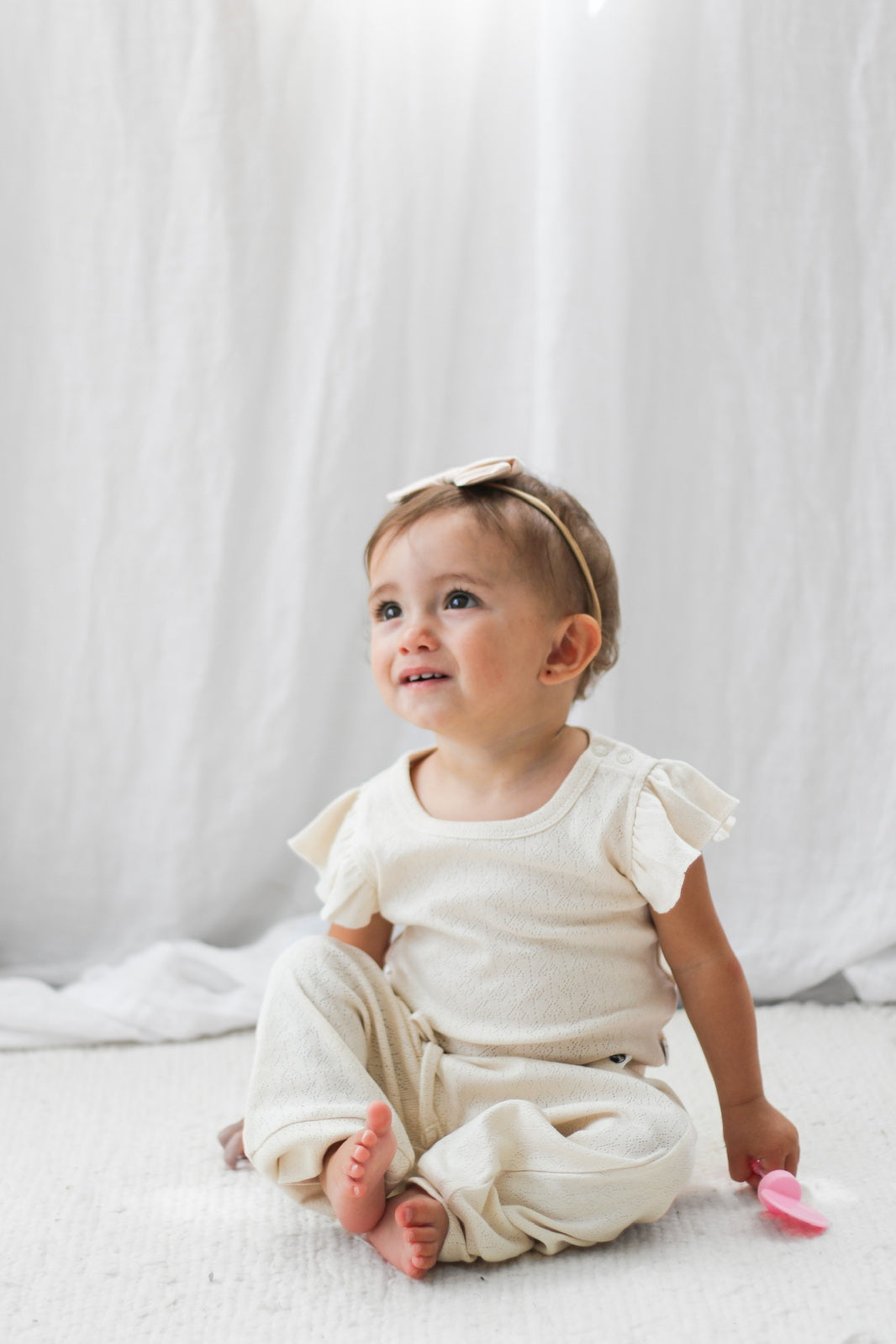 Baby sitting on white carpet in front of white curtain, posing while looking up and wearing in Pointelle Flutter Sleeve Bodysuit and pants in Natural, all designed by Burrow and Be
