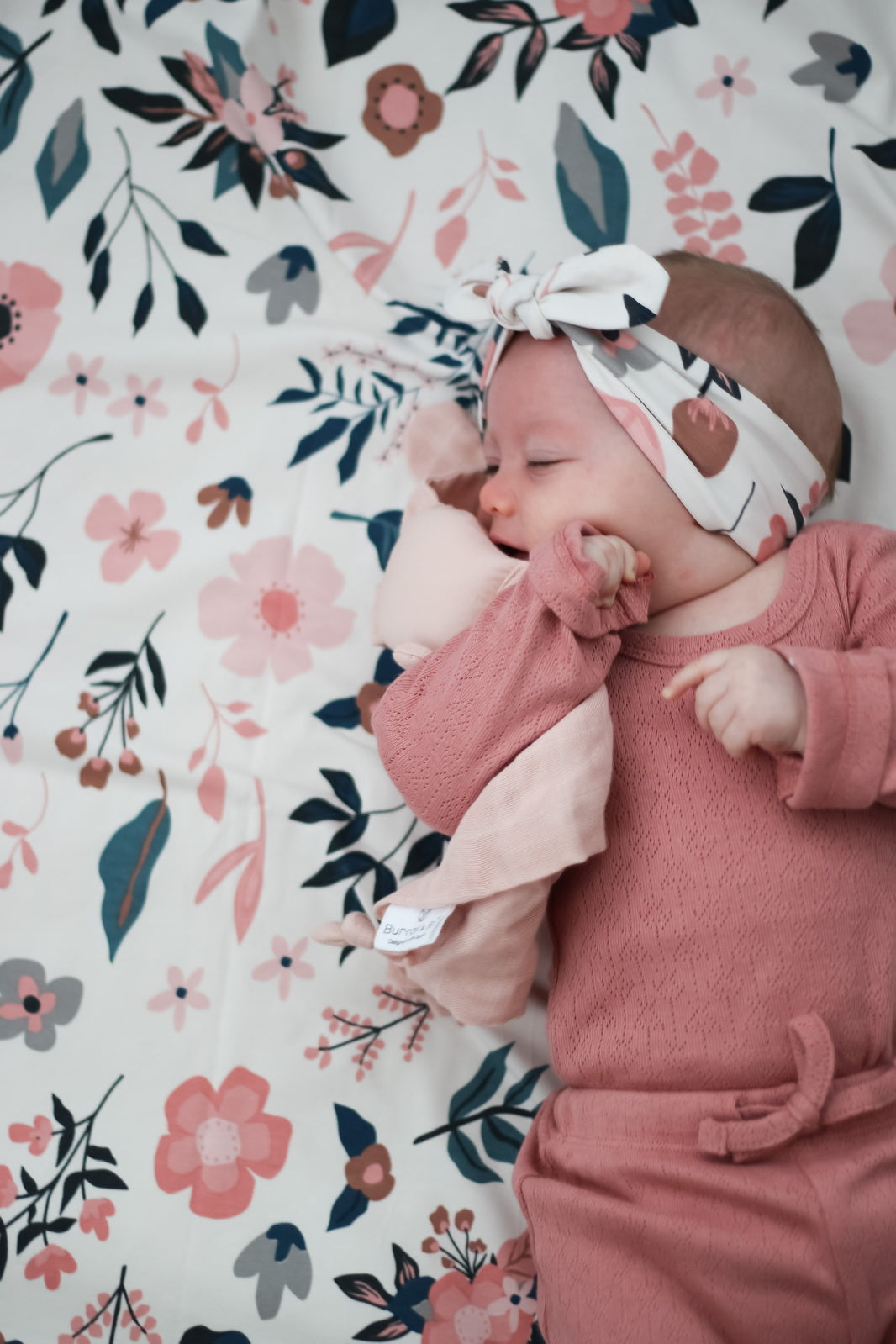 Baby laying on Pink Clementine Bed Sheets while wearing Pink Clementine Headband and Pointelle Long Sleeve Bodysuit and Baby Pants in Dusk, all designed by Burrow and Be