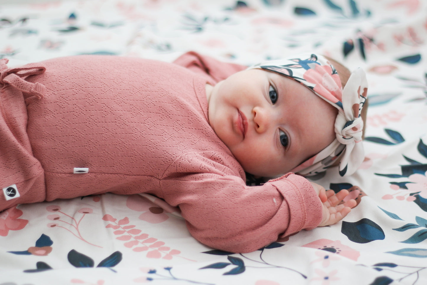 Baby laying on Pink Clementine Bed Sheets while wearing Pink Clementine Headband and Pointelle Long Sleeve Bodysuit and Baby Pants in Dusk, all designed by Burrow and Be