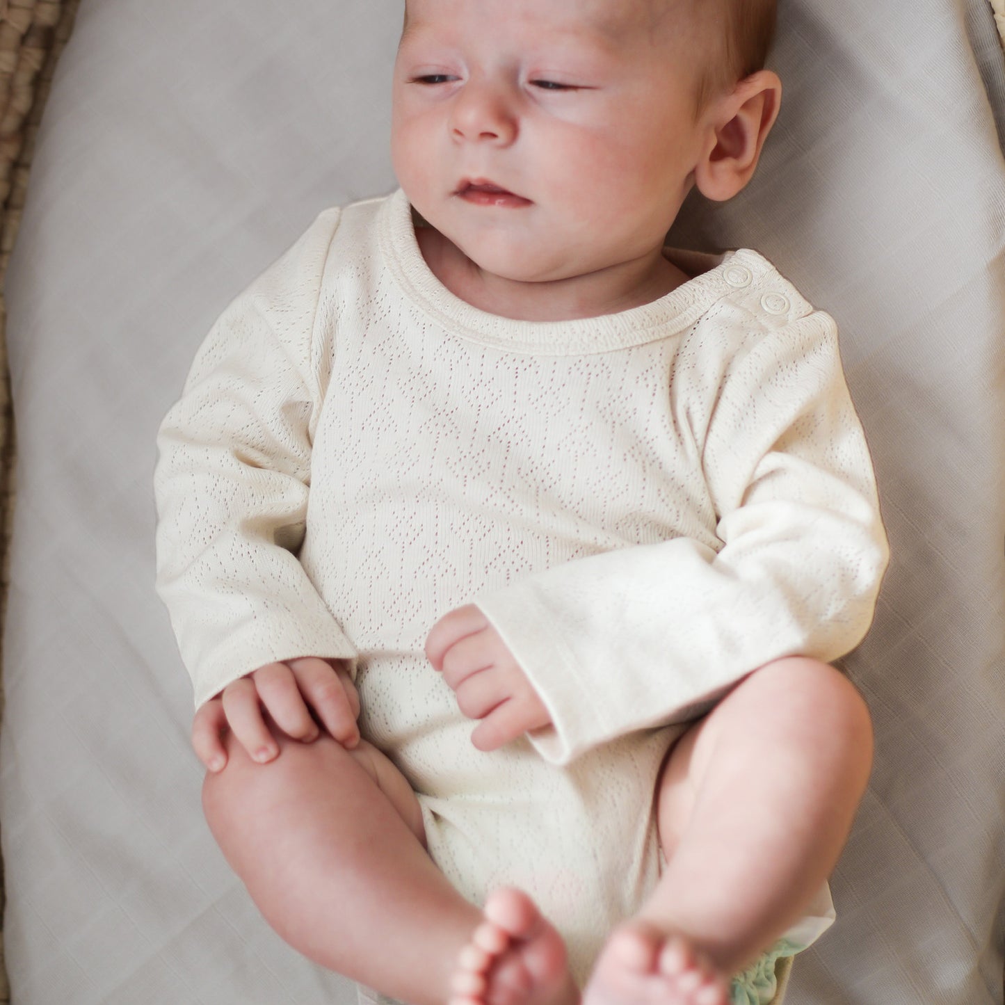 Close up of Baby laying in Moses Basket with Sage Sheets while wearing Pointelle Long Sleeve Bodysuit in Natural, all designed by Burrow and Be