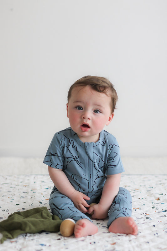 Baby sitting on bed in Burrow and Be Giant Bugs Zip Suit - Short Sleeve