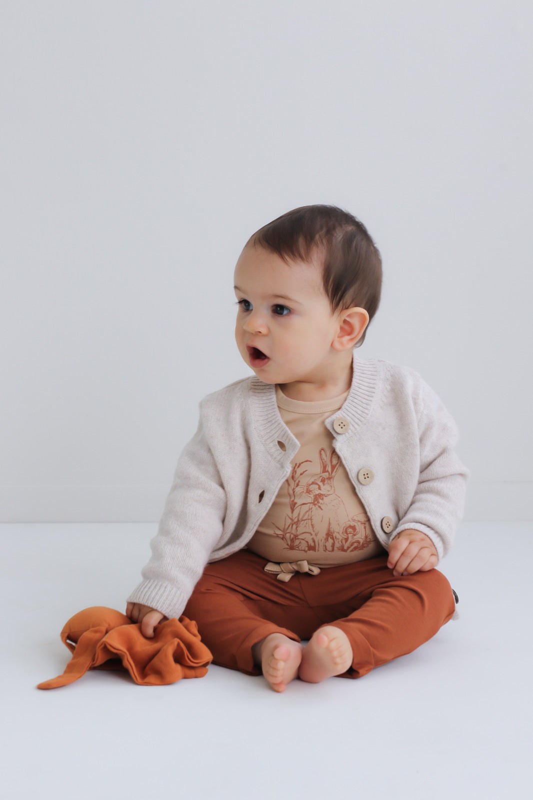 Baby boy sitting on wooden floor in front of a white background, wearing Marley Pants - Chestnut, designed by Burrow and Be
