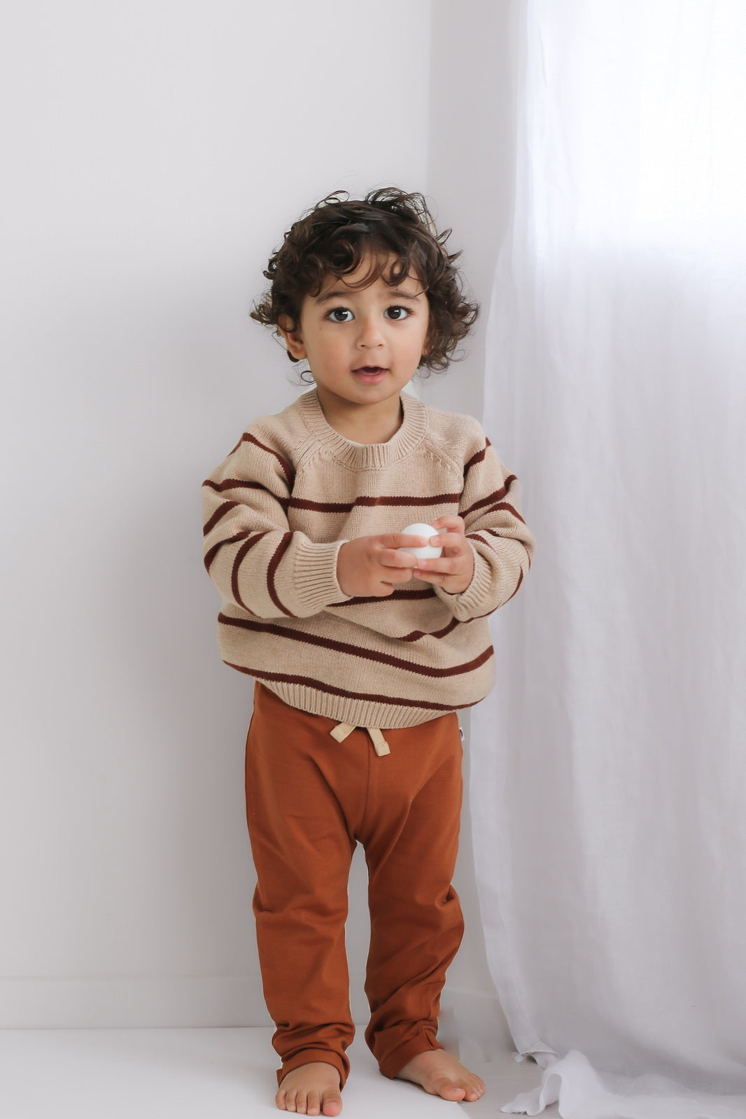 Boy standing in front of white curtains holding a ball and posing in Marley Pants - Chestnut, designed by Burrow and Be