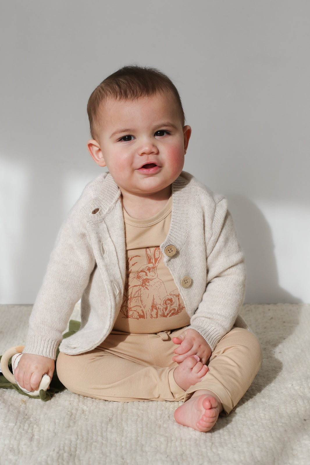 Baby boy sitting on carpet and posing in a Cardigan, Burrowers Bodysuit and Marley Pants - Latte, all designed by Burrow and Be