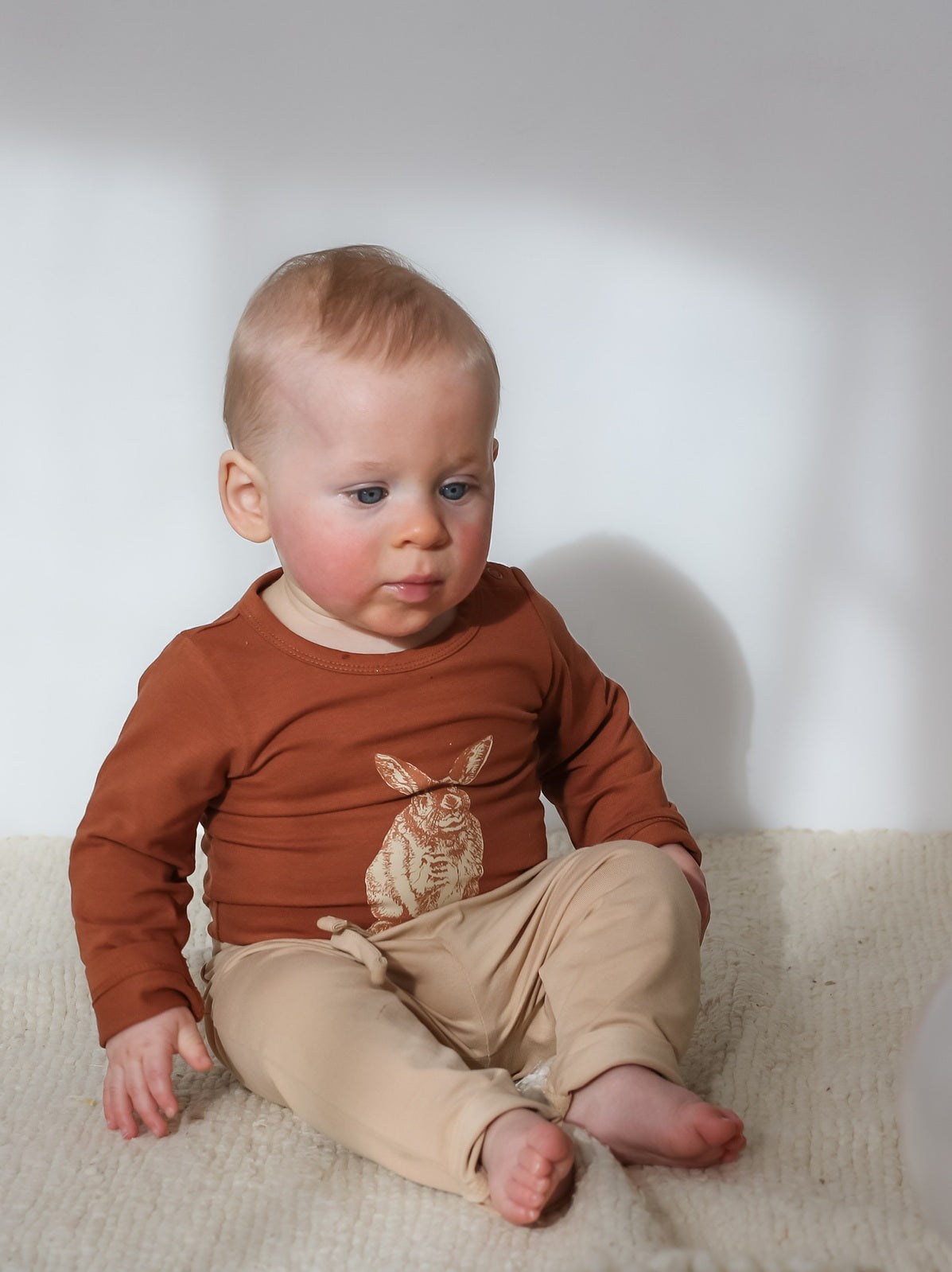 Baby sitting on a carpet in front of white background, wearing Burrowers Long Sleeve Bodysuit in Chestnut and Marley Pants - Latte, designed by Burrow and Be