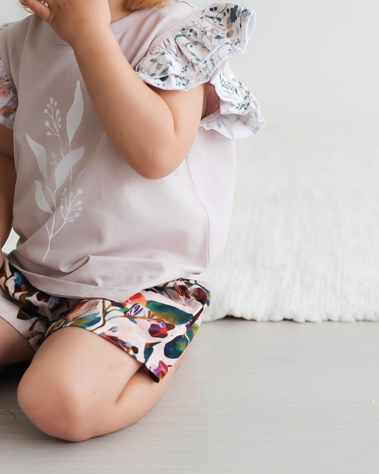Child kneeling on ground with white curtain background while wearing Wildflower/Lavender Flutter T-shirt and Garden Party Lyla Shorts by Burrow and Be