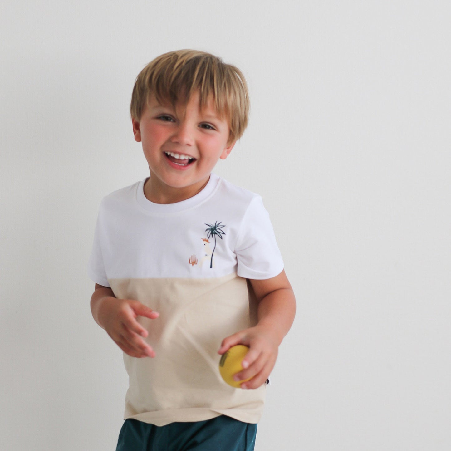 Boy holding a ball in hand and standing against a white wall while wearing Jungle Stamp Block T-Shirt, designed by Burrow and Be